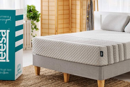 The Leesa Hybrid mattress sitting on a bed frame in a bedroom next to the box it came in. Leesa mattresses are up to $500 of for President's Day 2022.