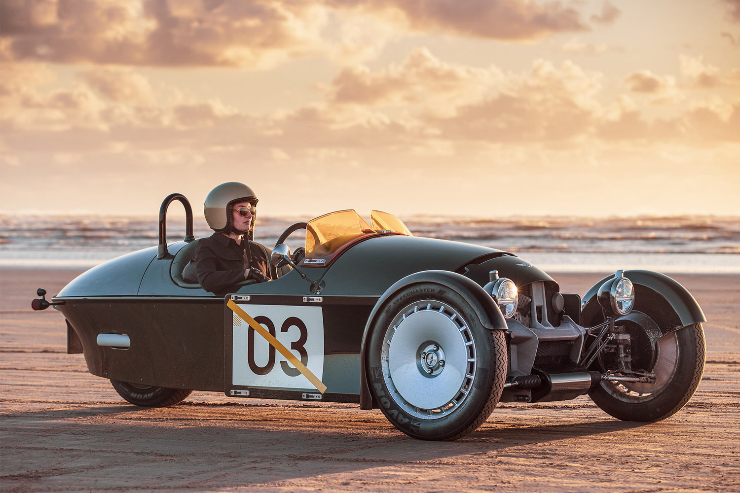 The new Morgan Super 3 in a green livery with the number 03 on the side. A helmeted passenger is seen sitting in the three-wheeled car on the sand.