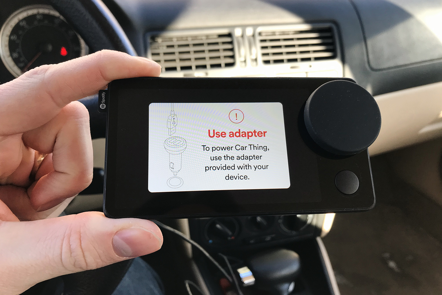 A hand holding up the Spotify Car Thing in a car, the screen showing an error that the device cannot connect to other 12-volt adapters other than the one included in the box