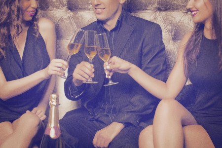 Image shows a man sitting on a couch surrounded by two women, all drinking champagne