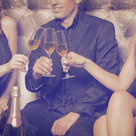 Image shows a man sitting on a couch surrounded by two women, all drinking champagne