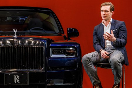Alex Innes, head of the Coachbuild Design team at Rolls-Royce Motor Cars, sitting next to the automaker's Cullinan SUV against a red background