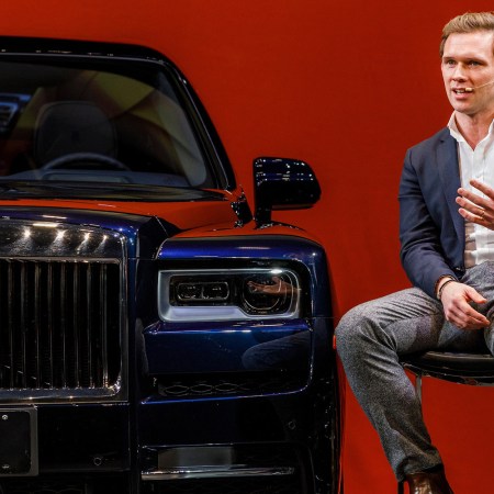 Alex Innes, head of the Coachbuild Design team at Rolls-Royce Motor Cars, sitting next to the automaker's Cullinan SUV against a red background