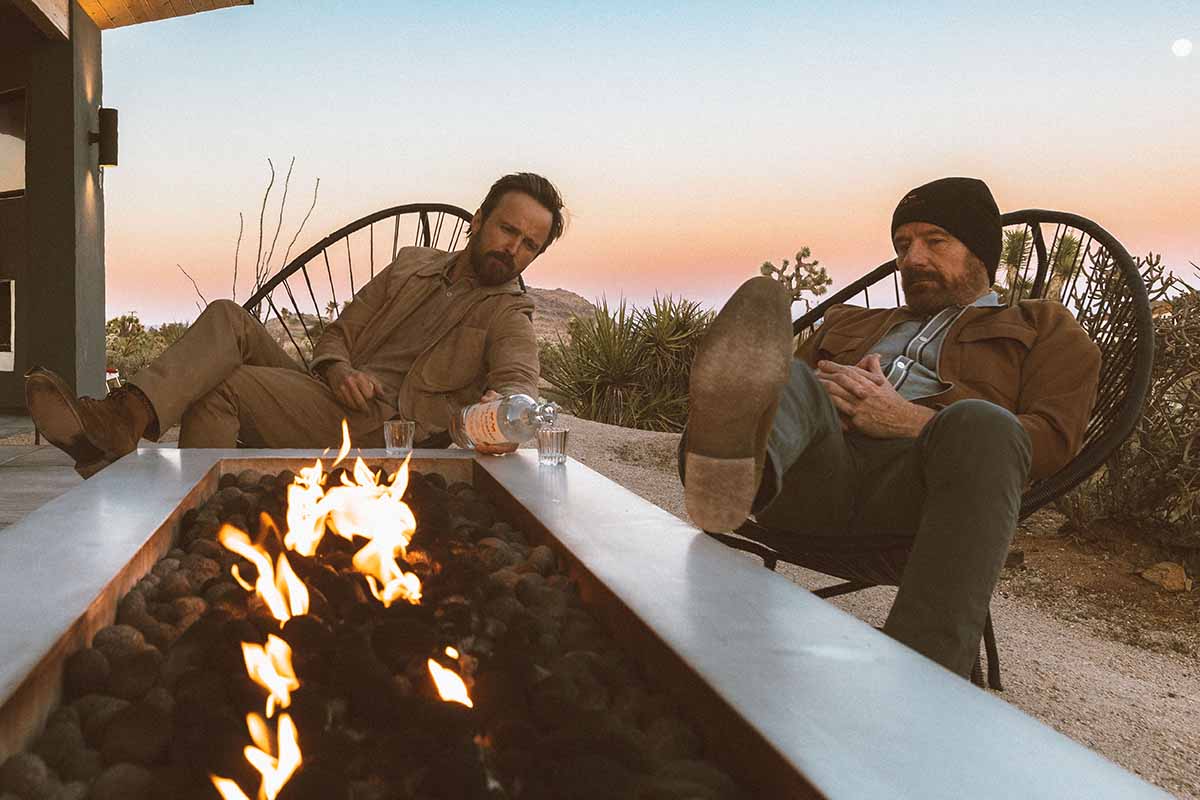Dos Hombres mezcal owners Aaron Paul and Bryan Cranston drinking mezcal on a porch in front of a fire pit