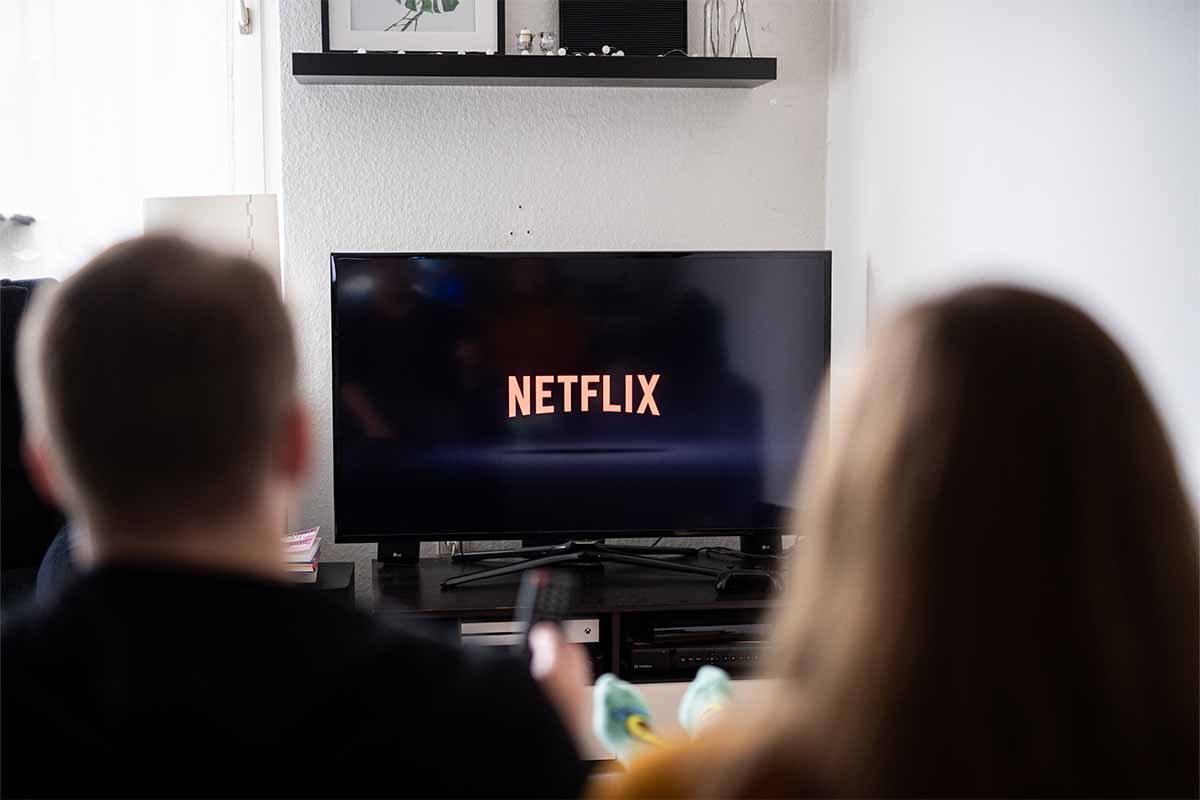 A couple sits in front of a television with the Netflix logo on it. Netflix may charge more if you share passwords "between households"