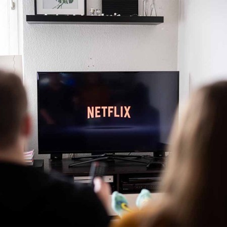 A couple sits in front of a television with the Netflix logo on it. Netflix may charge more if you share passwords "between households"