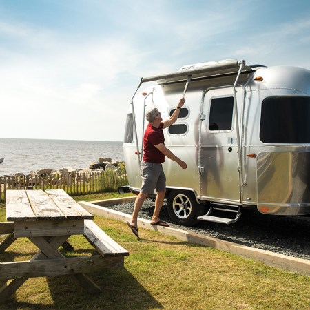 A man setting up his aluminum Airstream travel trailer next to a picnic table overlooking the ocean. According to new financial results, RV backlogs are still growing two years into the pandemic.
