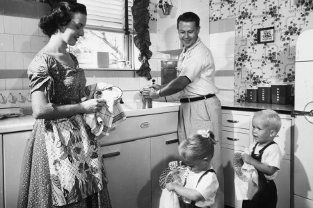 Black and white photo from the 1950s shows a mother and father receive help from their young children while washing and drying dishes in their kitchen.