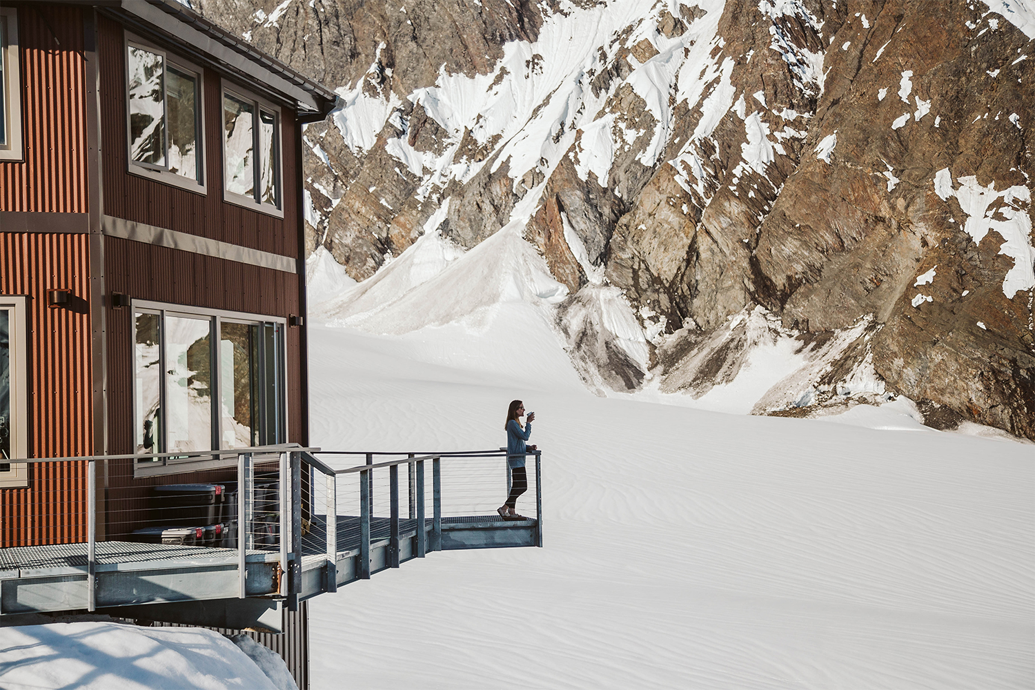 A woman drinking coffee on the balcony at Sheldon Chalet, Alaska, a remote, luxury destination