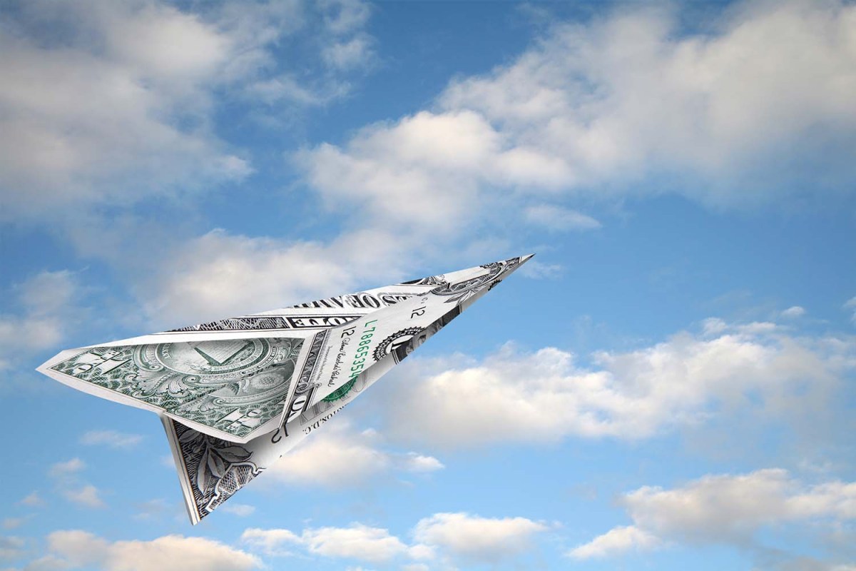 A folded up dollar bill in the shape of a paper airplane with the sky in the background. We look at the cities that have seen the steepest hikes in airfare in 2022.
