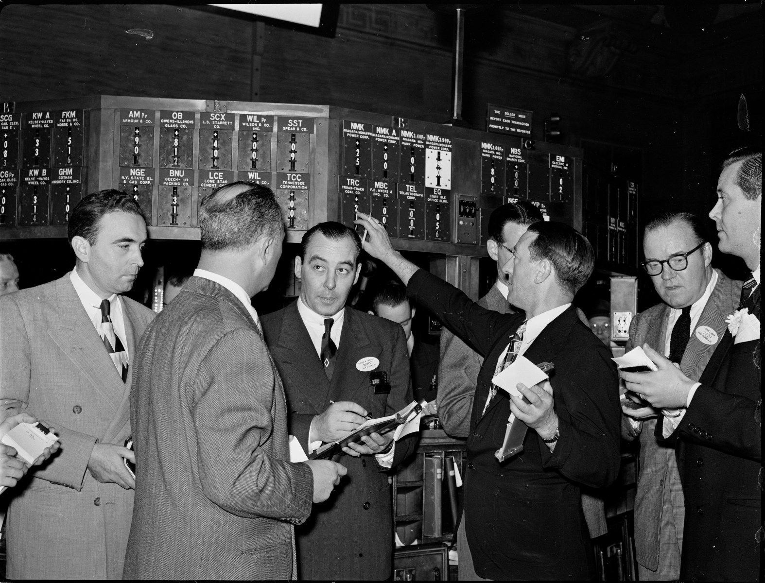 Here are some brokers at a trading post. These giant trading posts were all over the gigantic trading floor of the New York Stock Exchange. The guy on the right is actually physically rolling the numbers that would display the sale price of a particular stock. The guy on the left with the big notebook was a specialist. His job was to take down a stock trade in that giant notebook. It was a very labor-intensive world and there were a lot of complicated steps to get through.