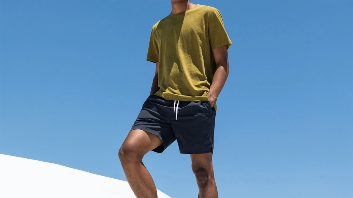 a model standing in blue everlane swim shorts and a yellow tee against a sky blue background