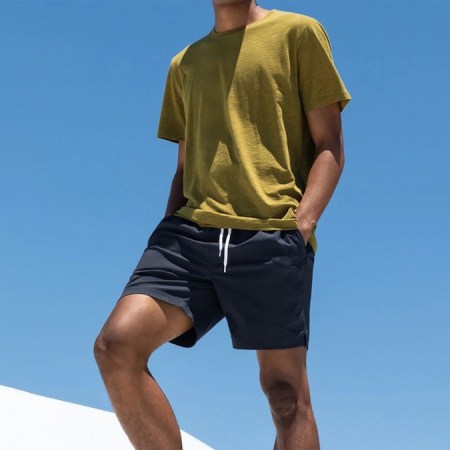 a model standing in blue everlane swim shorts and a yellow tee against a sky blue background