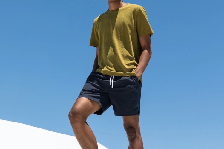 a model standing in blue everlane swim shorts and a yellow tee against a sky blue background