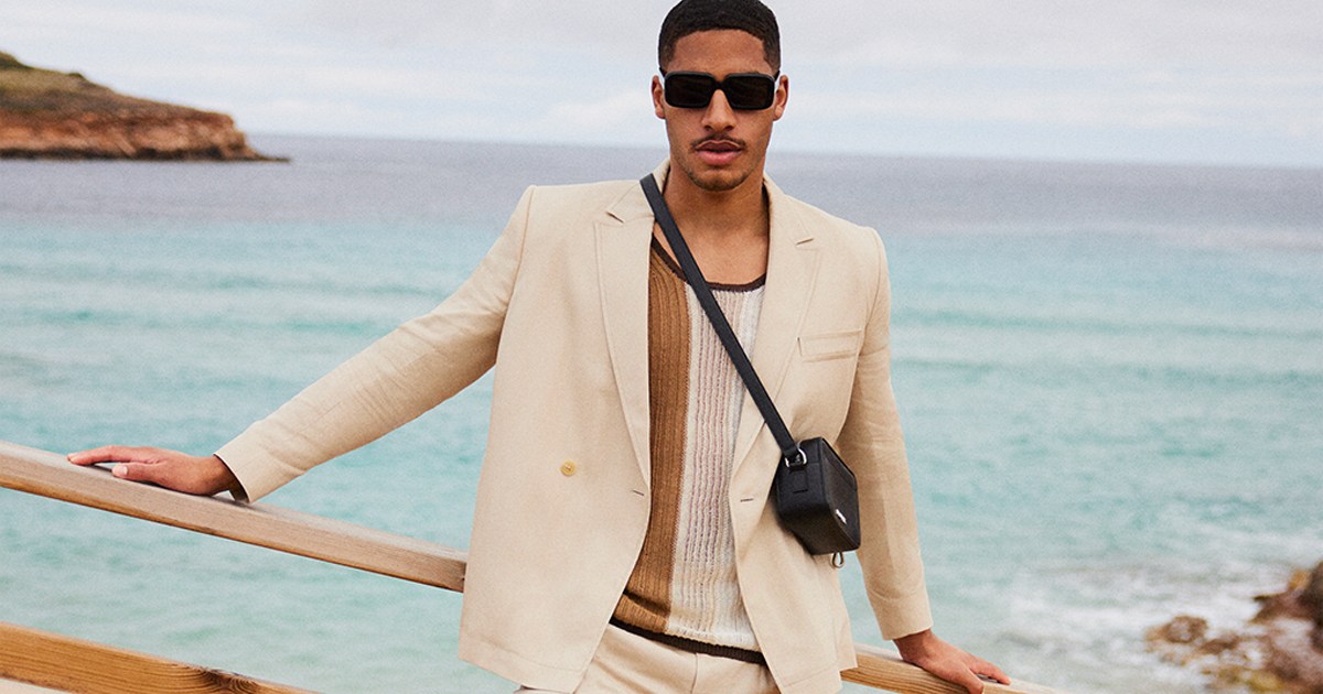 a model in a white suit, shirt and black leather bag standing in front of the ocean, courtesy of Mytheresa