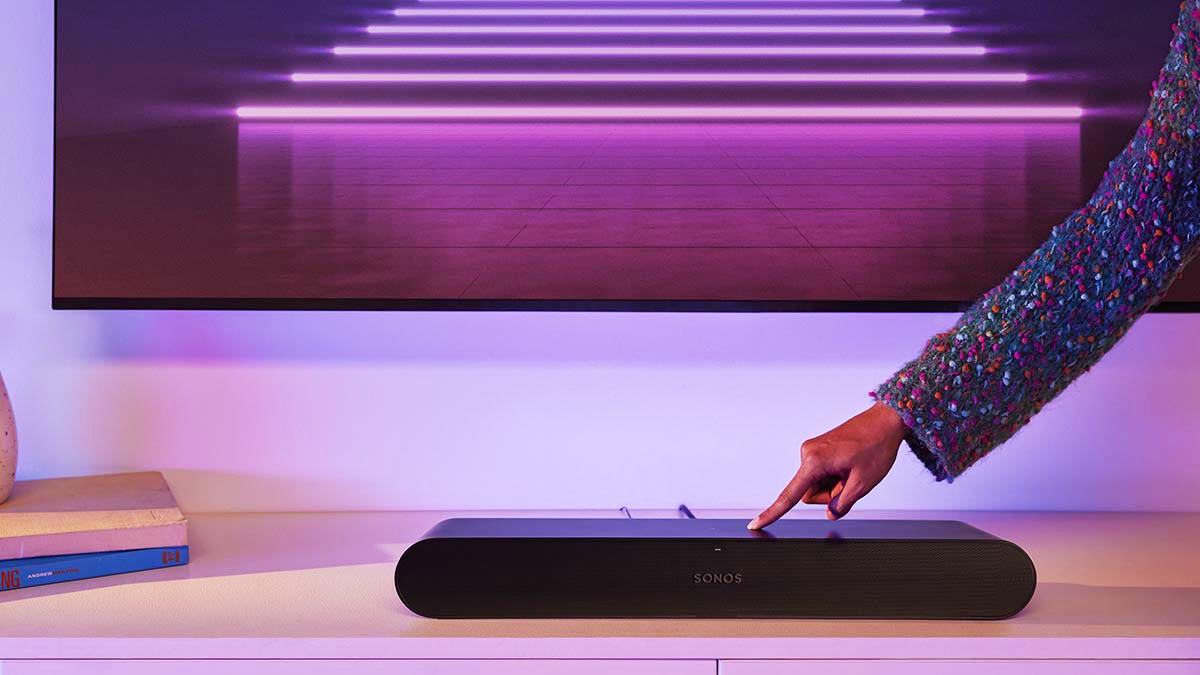 A Sonos Ray soundbar on a TV stand, with a hand adjusting the volume. The Ray is part of a new Sonos lineup