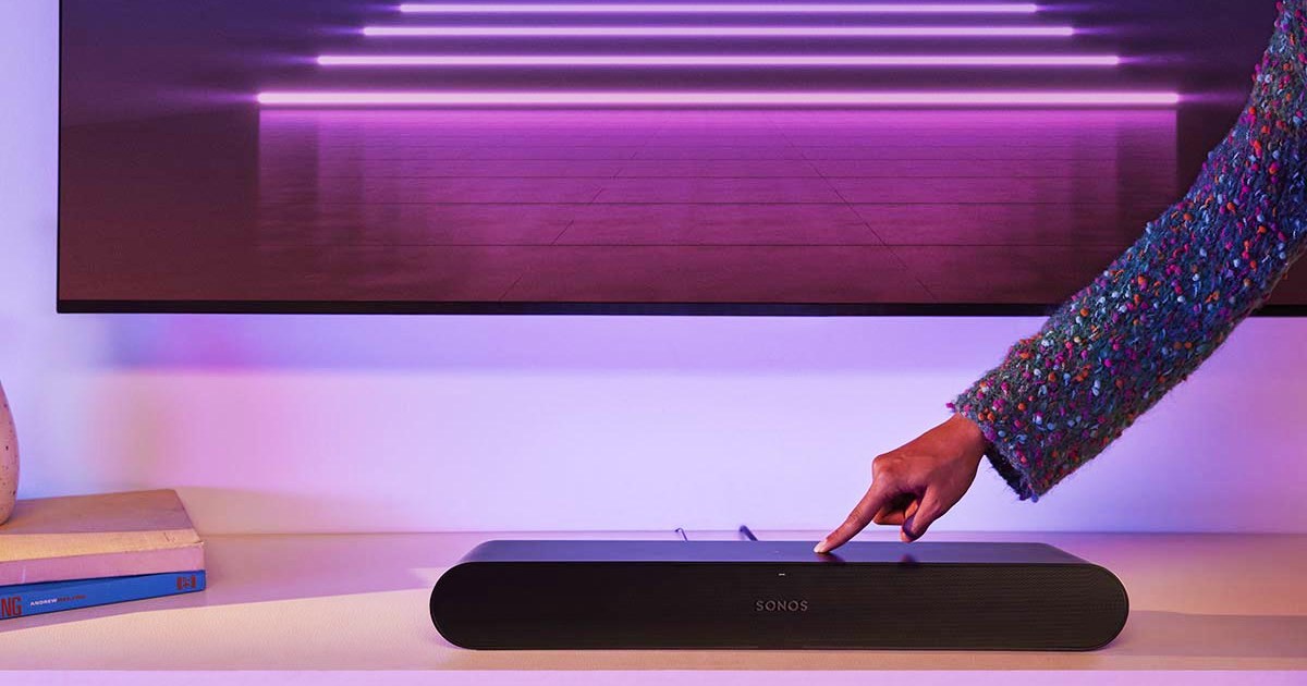 A Sonos Ray soundbar on a TV stand, with a hand adjusting the volume. The Ray is part of a new Sonos lineup