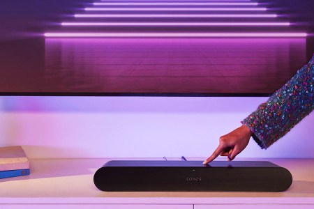 A Sonos Ray soundbar on a TV stand, with a hand adjusting the volume. The Ray is part of a new Sonos lineup