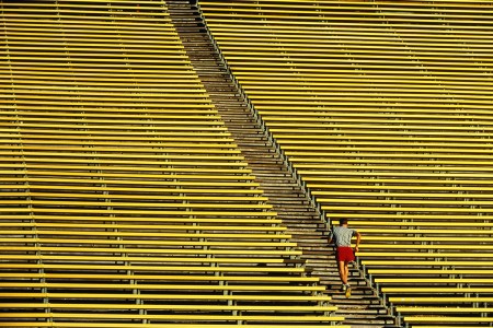 A man in red shorts and a grey shirt running up yellow steps in a football stadium.