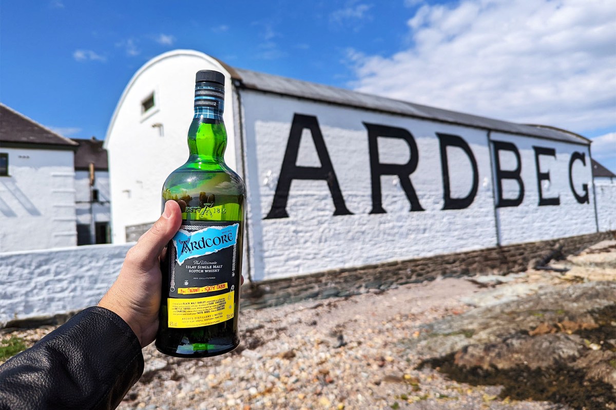 Writer Jake Emen's hand is seen in the photo holding up a bottle of Ardcore Scotch whisky in front of a building at the Ardbeg distillery on the island of Islay, Scotland