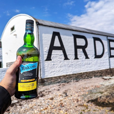 Writer Jake Emen's hand is seen in the photo holding up a bottle of Ardcore Scotch whisky in front of a building at the Ardbeg distillery on the island of Islay, Scotland