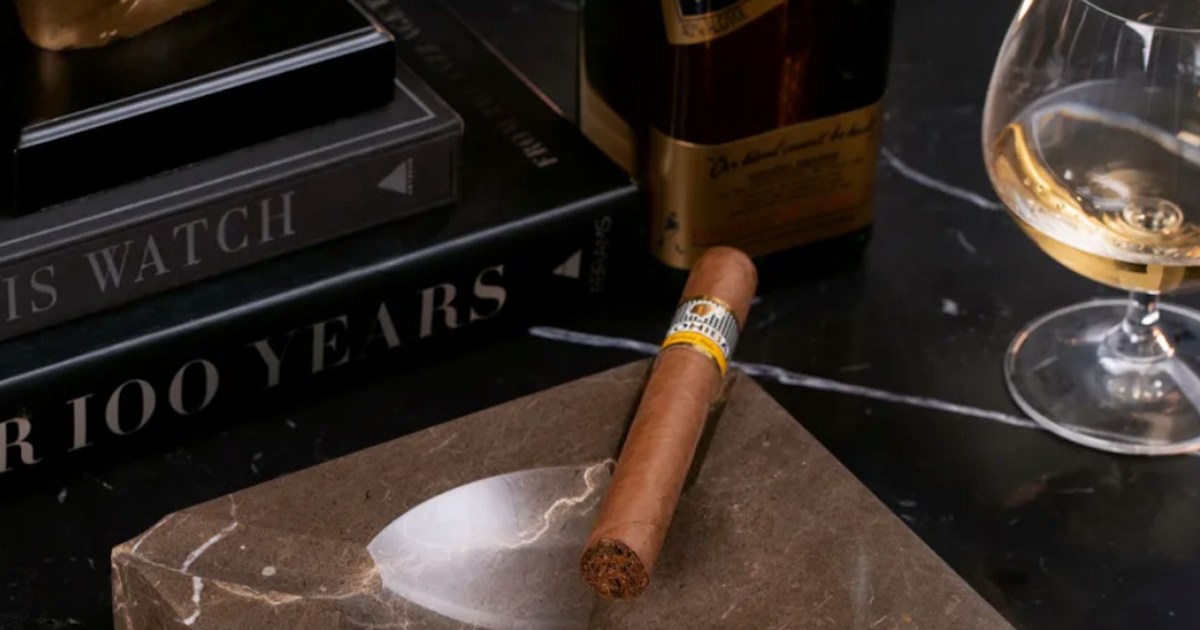 a stock shot of a cigar and ashtray, as well as books and whiskey, on a marble countertop