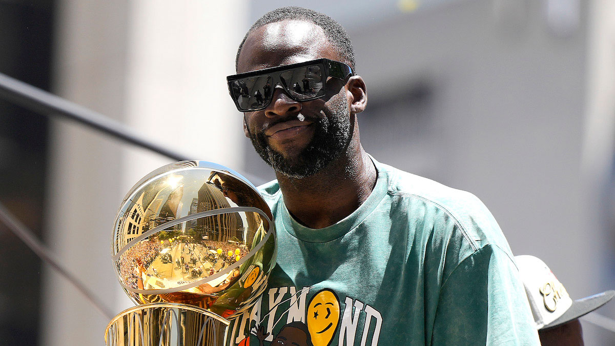 A photo of Golden State Warriors Forward Draymond Green wearing sunglasses and a green shirt and holding the 2022 NBA championship trophy