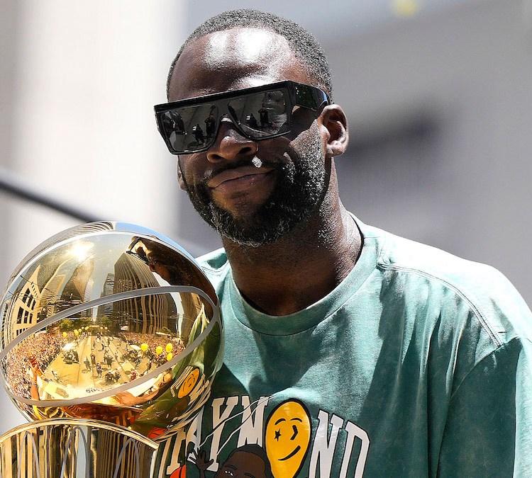 A photo of Golden State Warriors Forward Draymond Green wearing sunglasses and a green shirt and holding the 2022 NBA championship trophy