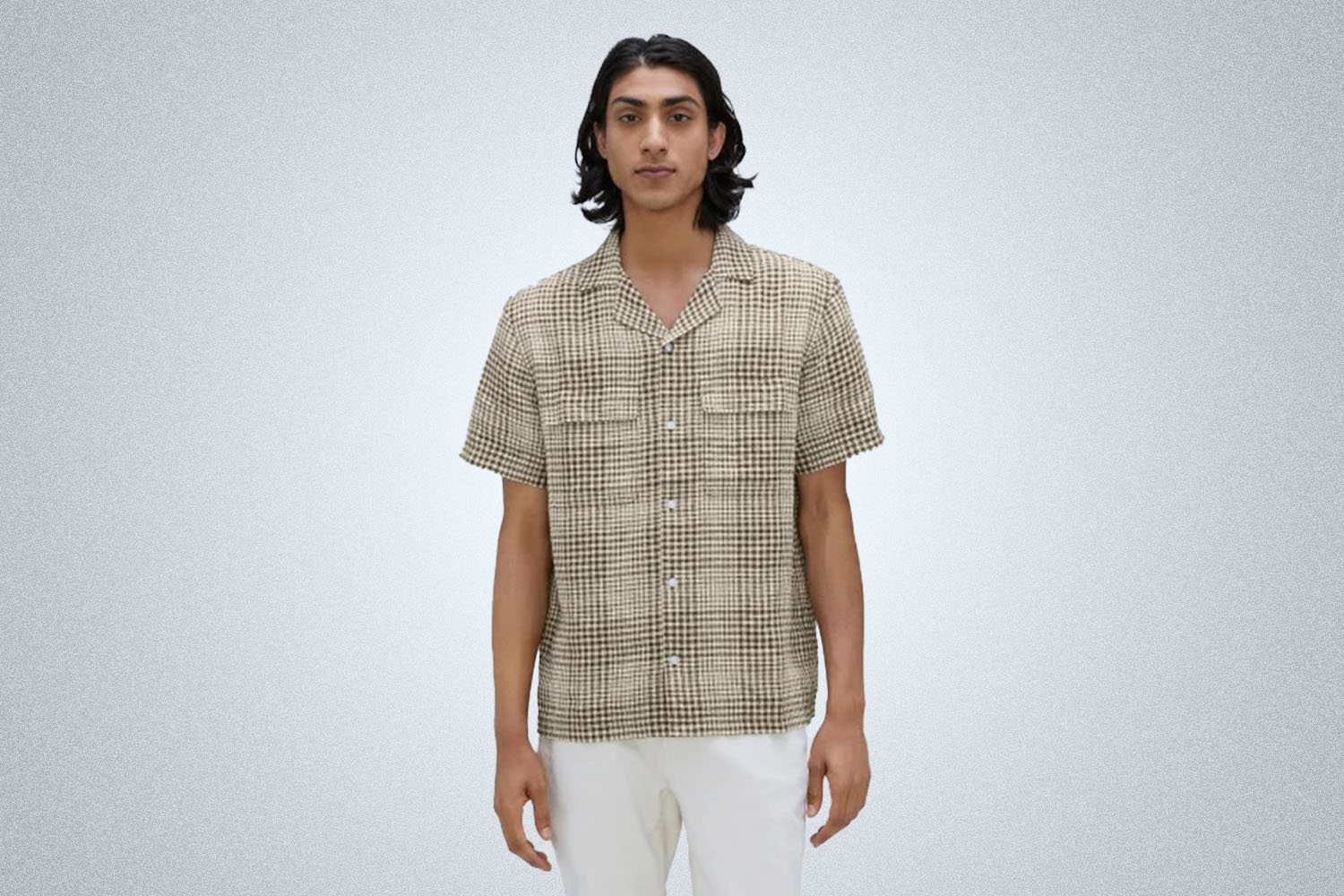 a model in a checked tan camp collar shirt and white pants from Everlane on a grey background