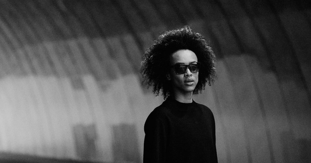 a black and white photo of a model in a black shirt standing in a tunnel wearing a pair of the District Vision x Reigning Champ eyewear collection