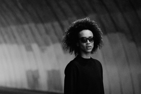 a black and white photo of a model in a black shirt standing in a tunnel wearing a pair of the District Vision x Reigning Champ eyewear collection