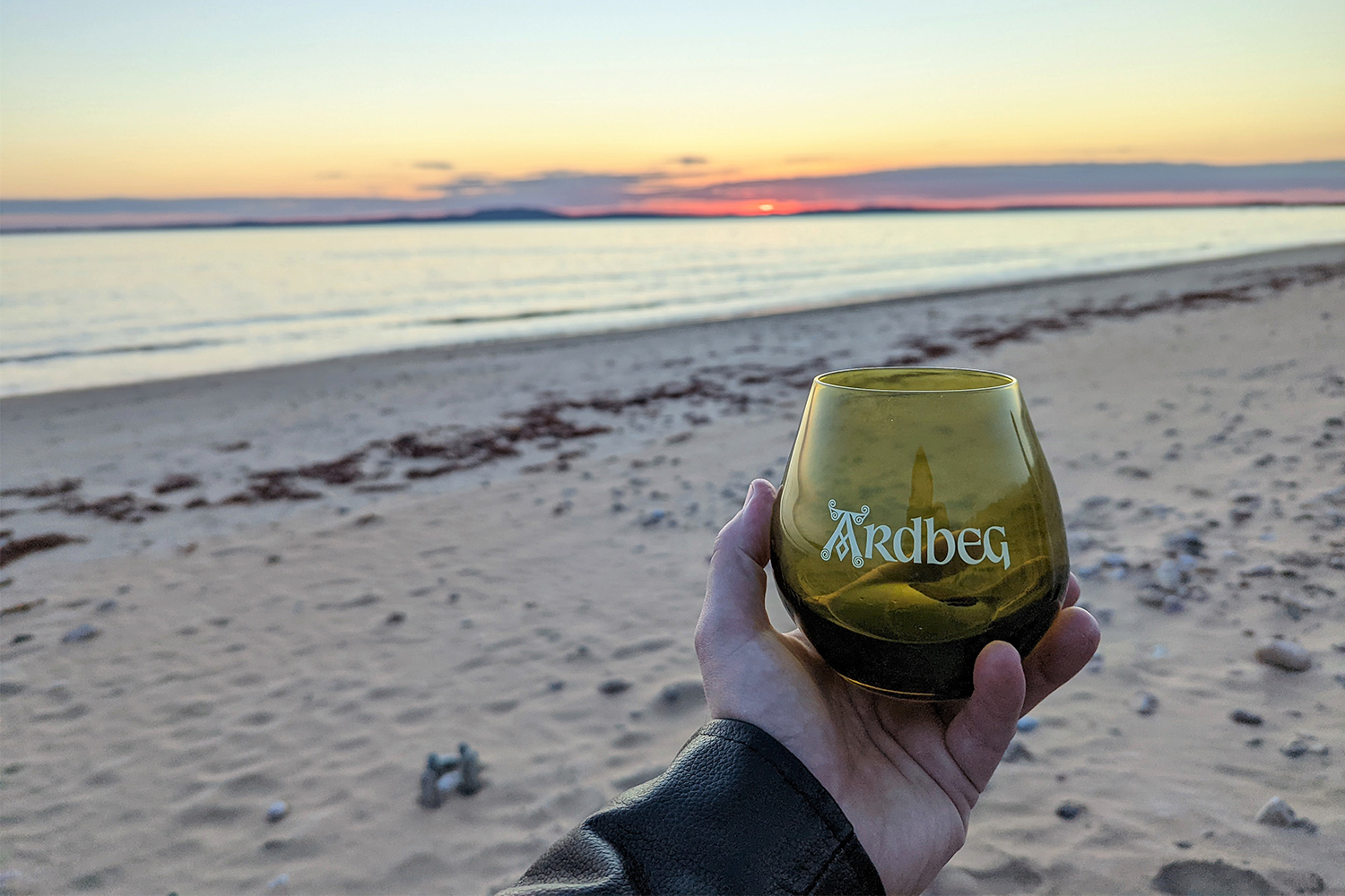 A hand holding a whiskey tasting glass on the beach in Islay, Scotland during the 2022 Island Feis of the Island 