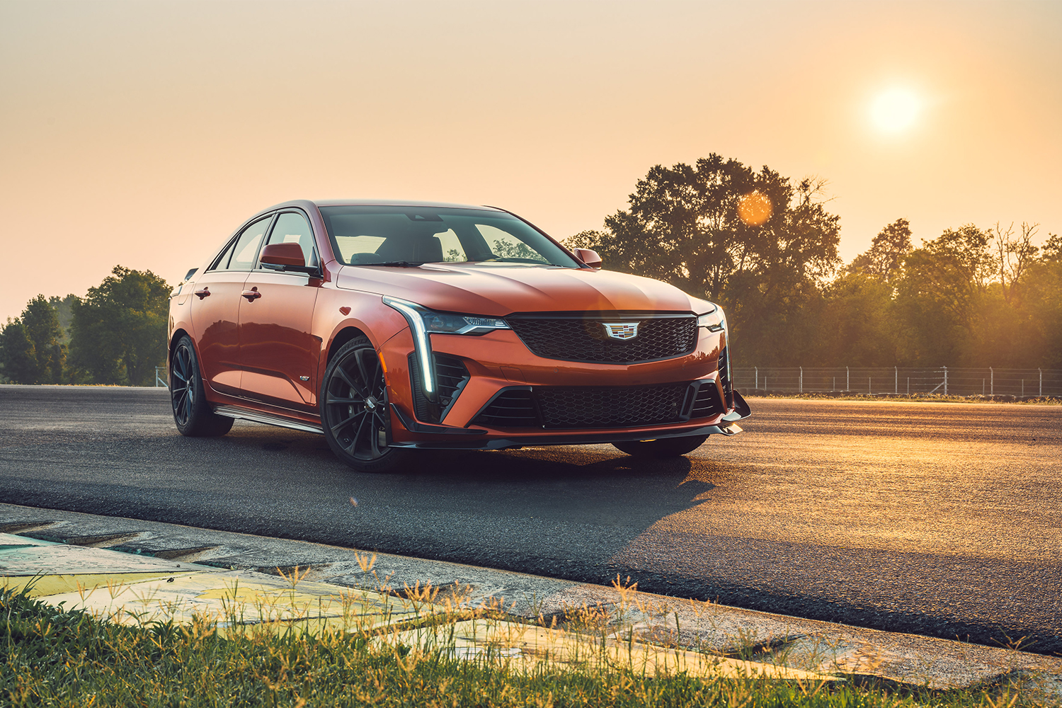 The Cadillac CT4-V Blackwing in orange sitting on a track with the sun setting in the background. We reviewed the 2022 V6 sport sedan and its sibling the CT5-V Blackwing.