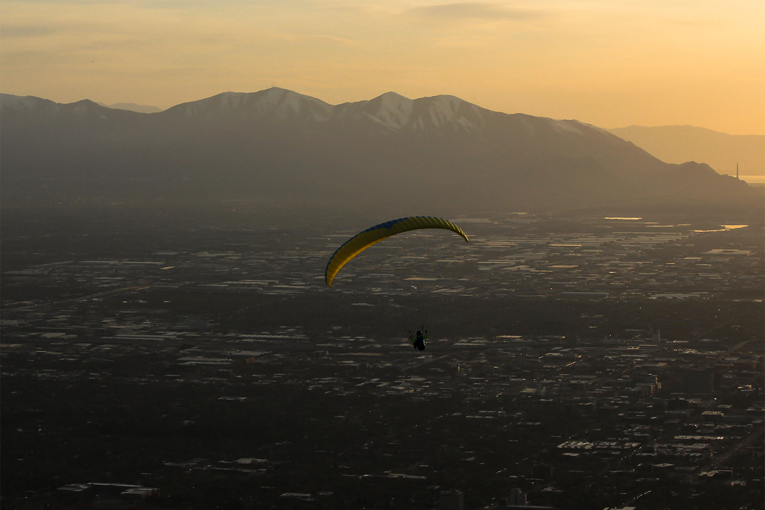 A paraglider flying over Salt Lake City, Utah, at sunset, with the mountains in the background under a yellow sky