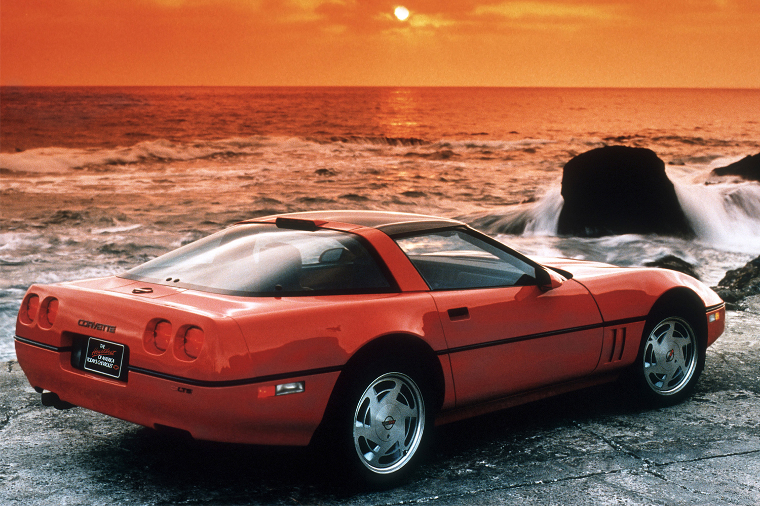 A red 1990 Chevrolet Corvette C4 sitting next to the ocean while the sun sets in an orange sky in the background