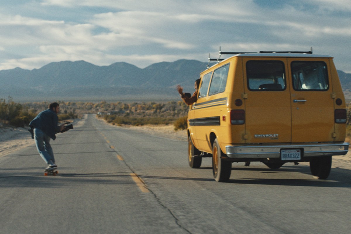 Man skateboards beside a moving yellow van on empty highway