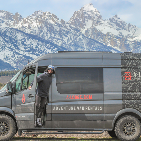 Writer and adventurer Joe Kanzangu stands in the door of his adventure van from A-Lodge with snow-capped mountain peaks in the background