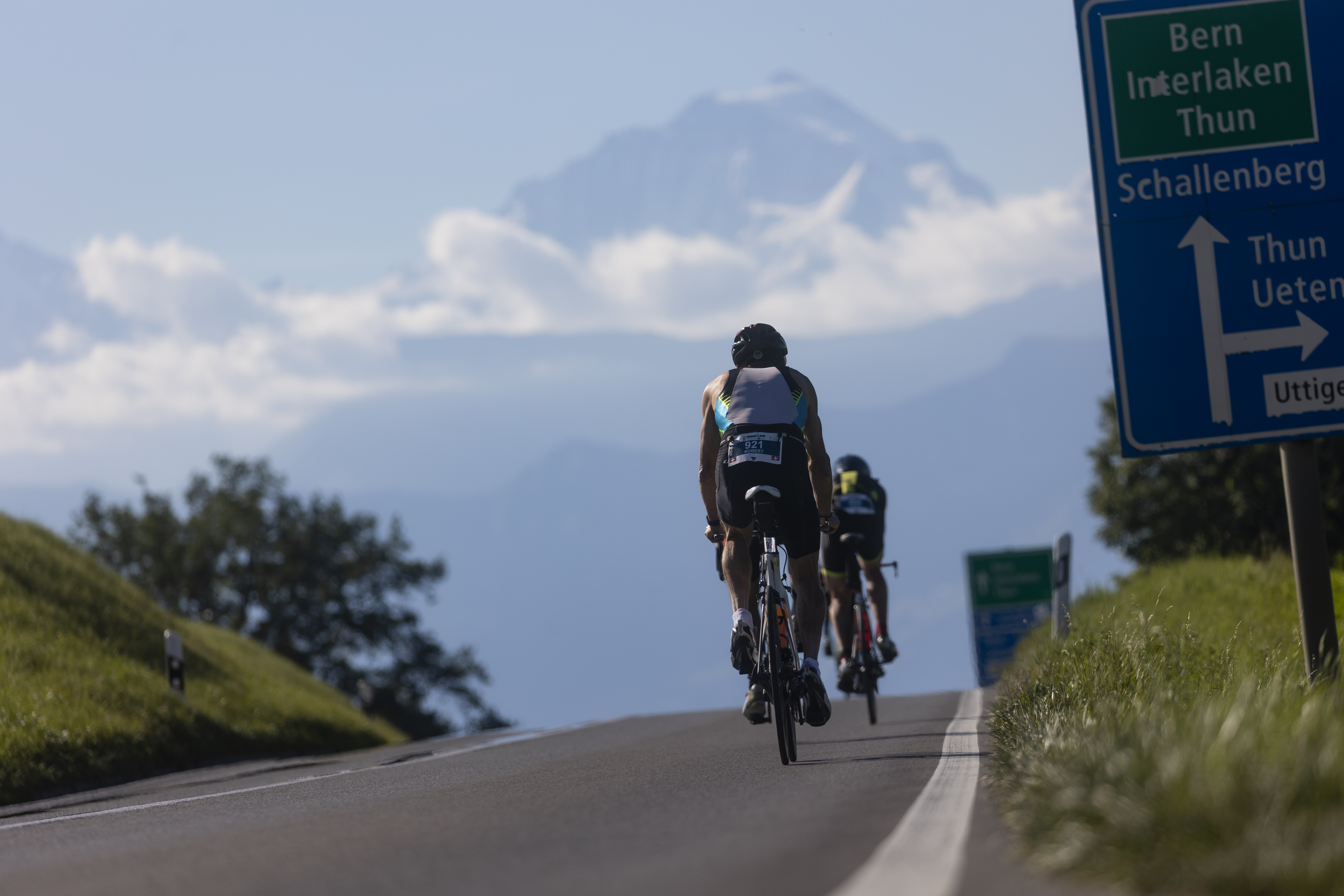 A man biking on a road in Switzerland with a mountain and clouds in the background