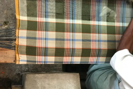 A green, red and beige madras pattern being made by an artisan