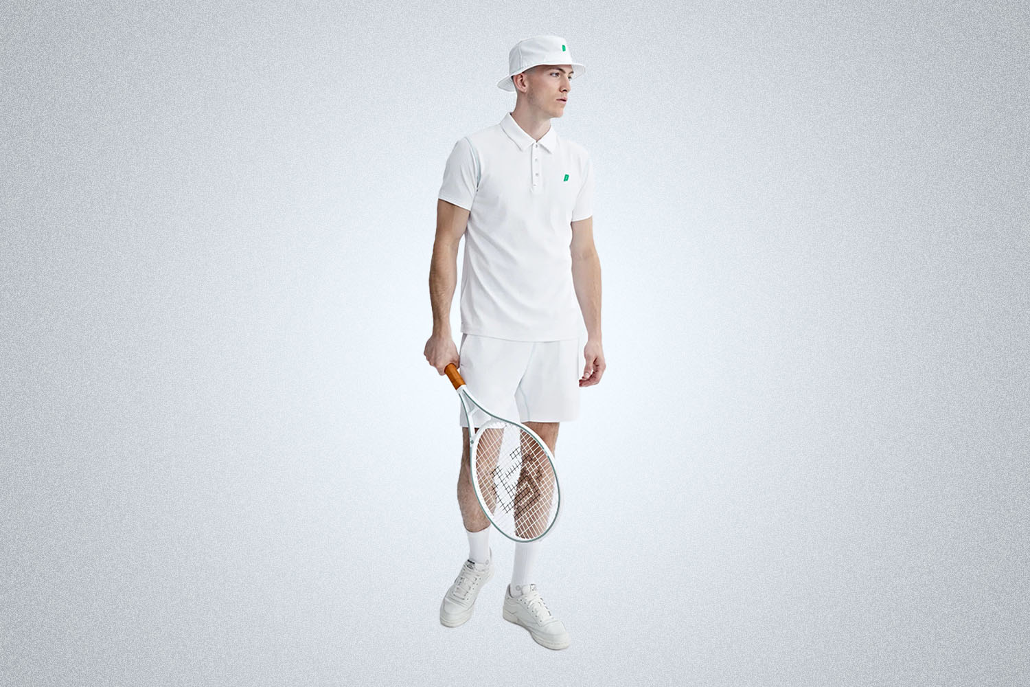 a model from the reigning champ x prince collection in all white, wearing a bucket hat, polo shirt, shorts and holding a tennis racket