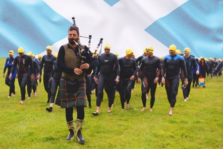 A group of athletes about to compete in the Great Kindrochit Quadrathlon are led to a lake by a bagpiper with a Scottish flag in the background.