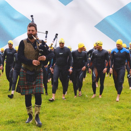 A group of athletes about to compete in the Great Kindrochit Quadrathlon are led to a lake by a bagpiper with a Scottish flag in the background.