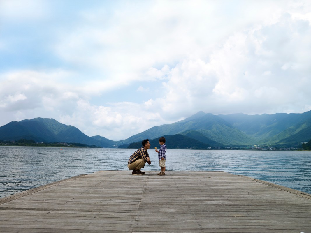 A father standing with his son on a pier in a lake surrounded by mountains.