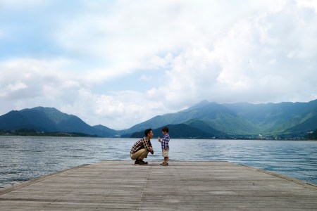 A father standing with his son on a pier in a lake surrounded by mountains.