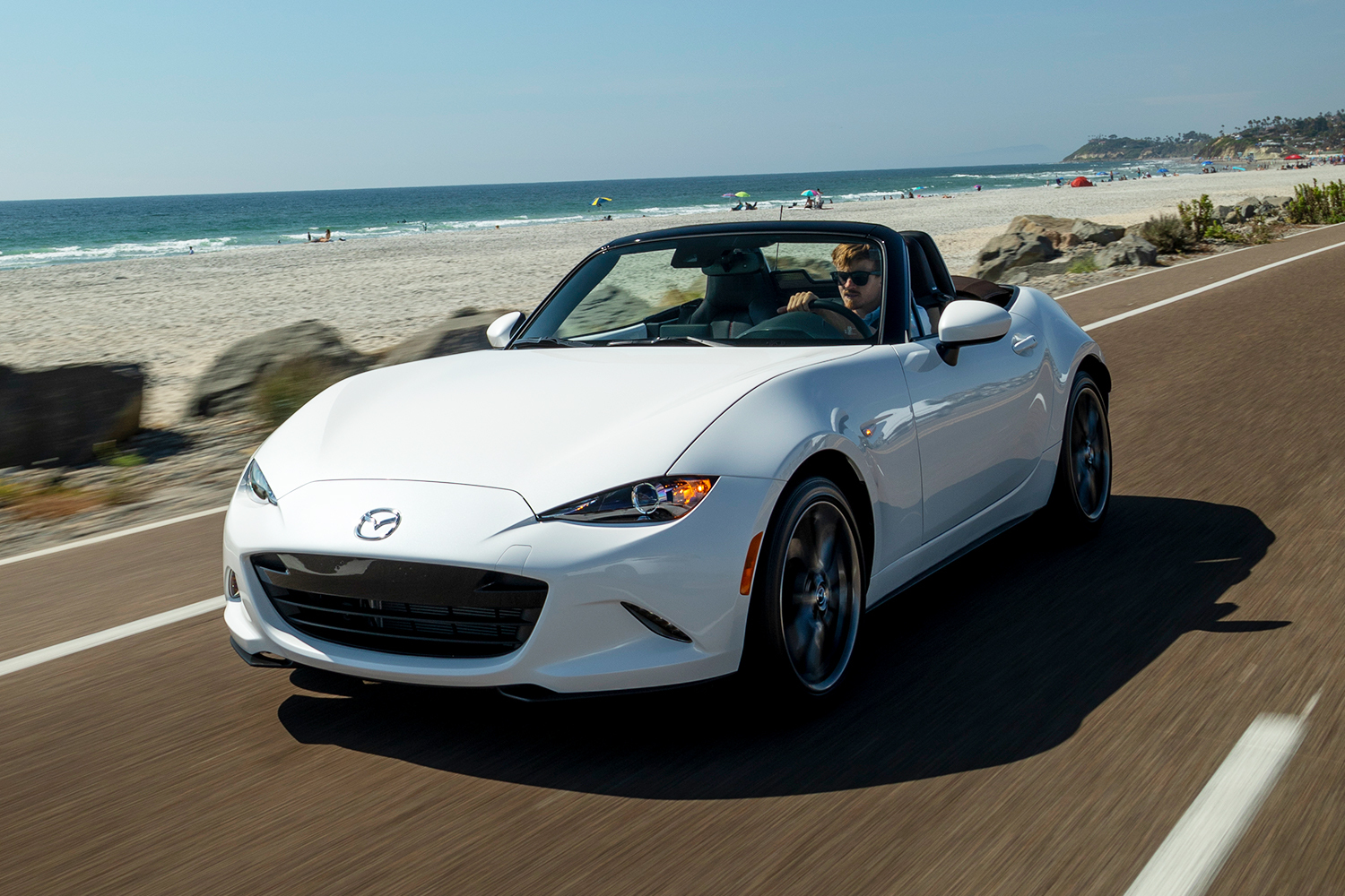 A man driving a white Mazda MX-5 Miata convertible next to an ocean beach
