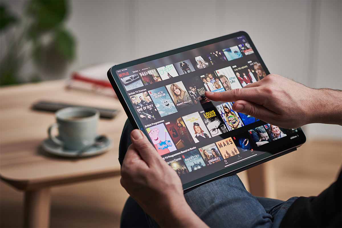 Detail of a mans hand scrolling through Netflix on an Apple iPad Pro. In certain countries, Netflix is cracking down on password sharing through a confusing new system.
