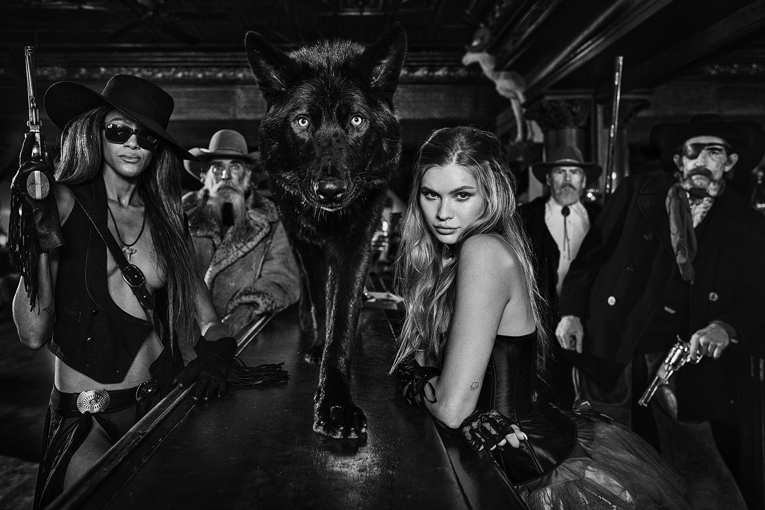 The photo "The Residents" by David Yarrow is a black and white Wild West image with a wolf on a bar and female and male actors dressed in cowboy attire in the background