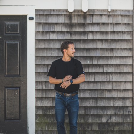 A man with a perfect tucked in black t-shirt standing in front of a grey wall.