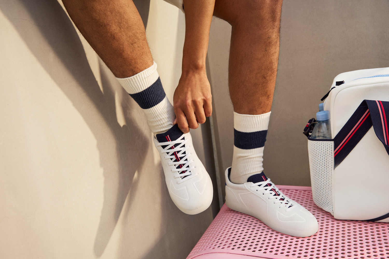 a model fixing a pair of white Rothy's x evian sneakers against a concrete and pink background