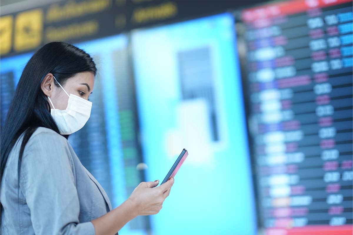 Woman wearing protective face mask checking flight schedule on mobile phone while standing in front of the arrival departure board. A new Twitter scam has fraudulent airline accounts attempting to take money from passengers